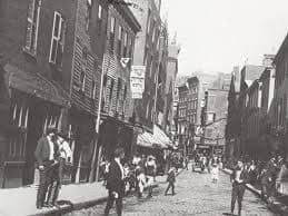 A black and white photograph of Boston's North End in the 1930s. Narrow, crowded Italian American street scene with tenements, laundry lines, and men standing on a corner. Evocative, documentary feel.