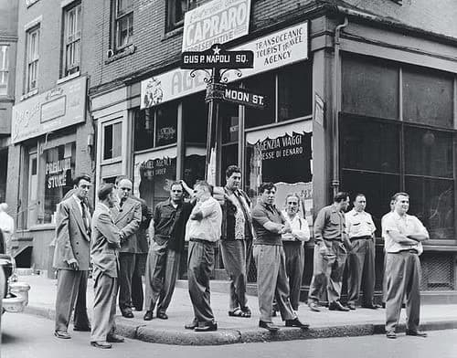 A black and white photograph of a North End / Boston street corner in the mid-1930s showing groups of men gathered, standing around, talking. Daily street life, not posed. Documentary feel.